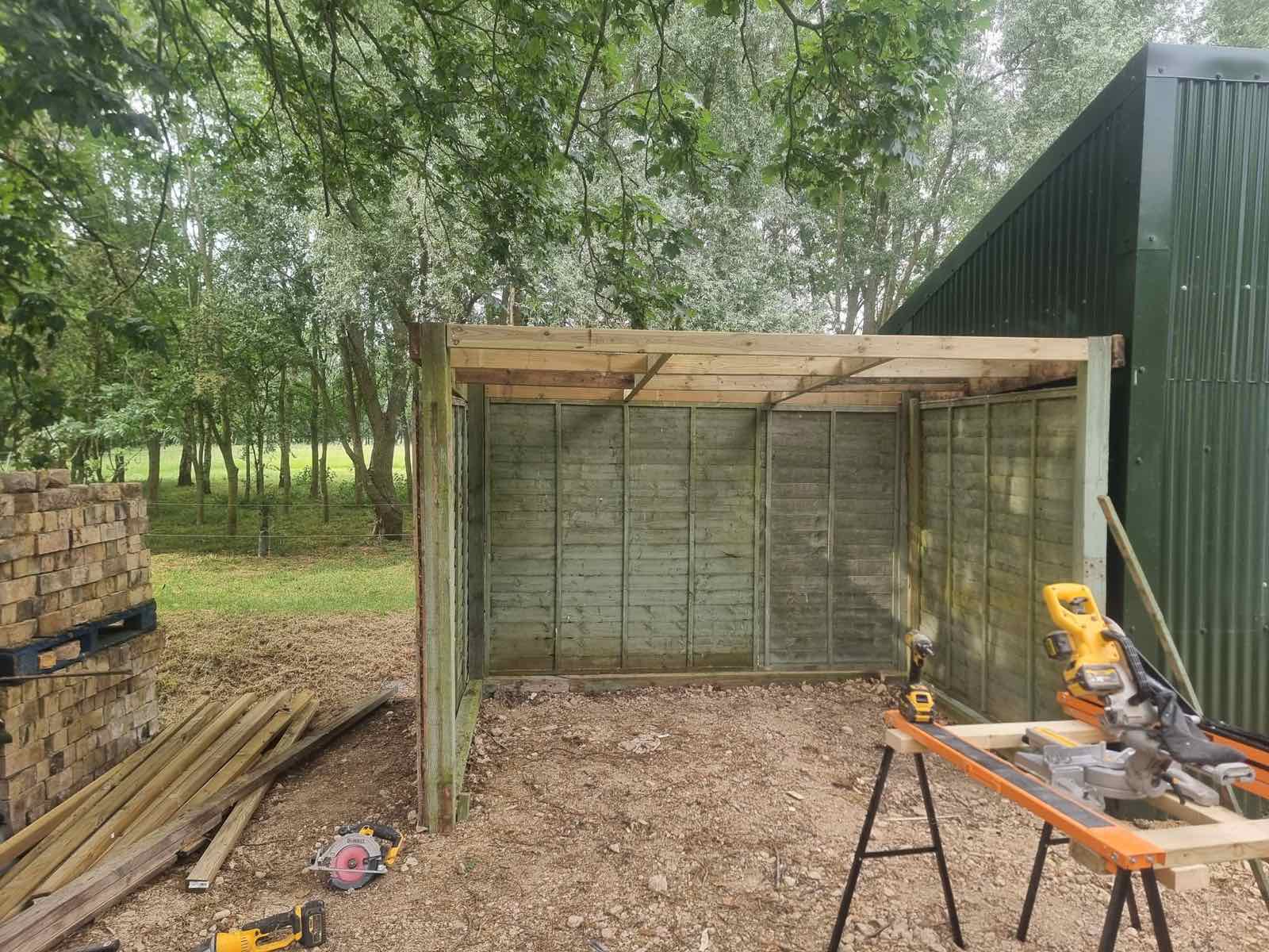 Timber-framed lean-to store under construction beside a garden outbuilding